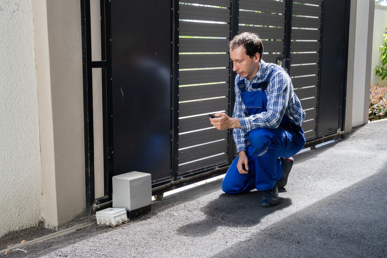 Man in blue overalls kneels, operating an automatic gate with a remote control on a driveway.