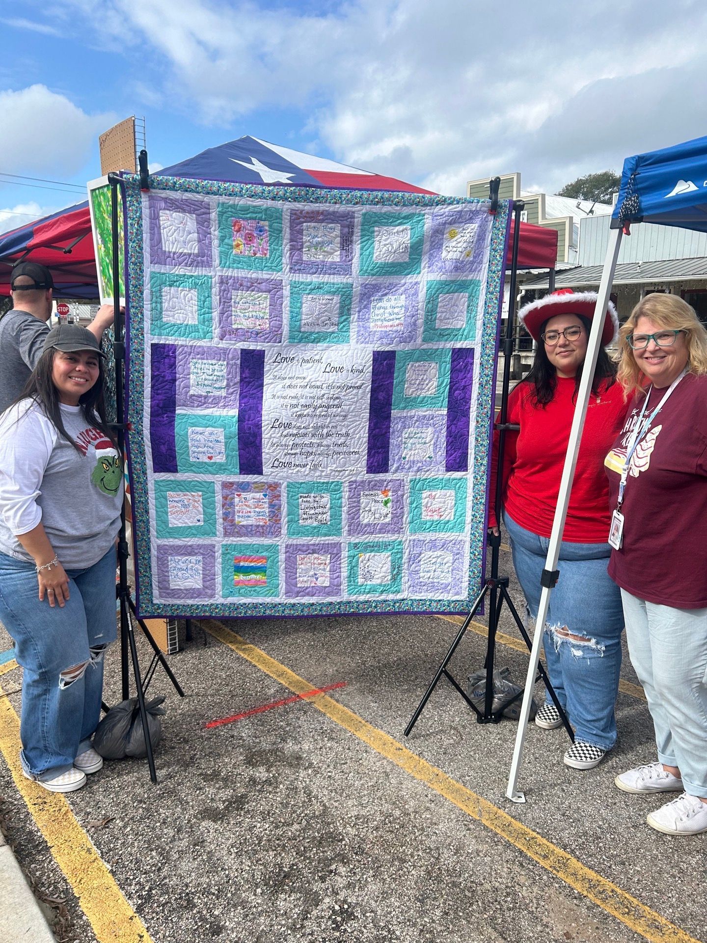 Three people stand by a quilt, smiling. The quilt is displayed outdoors on a stand.