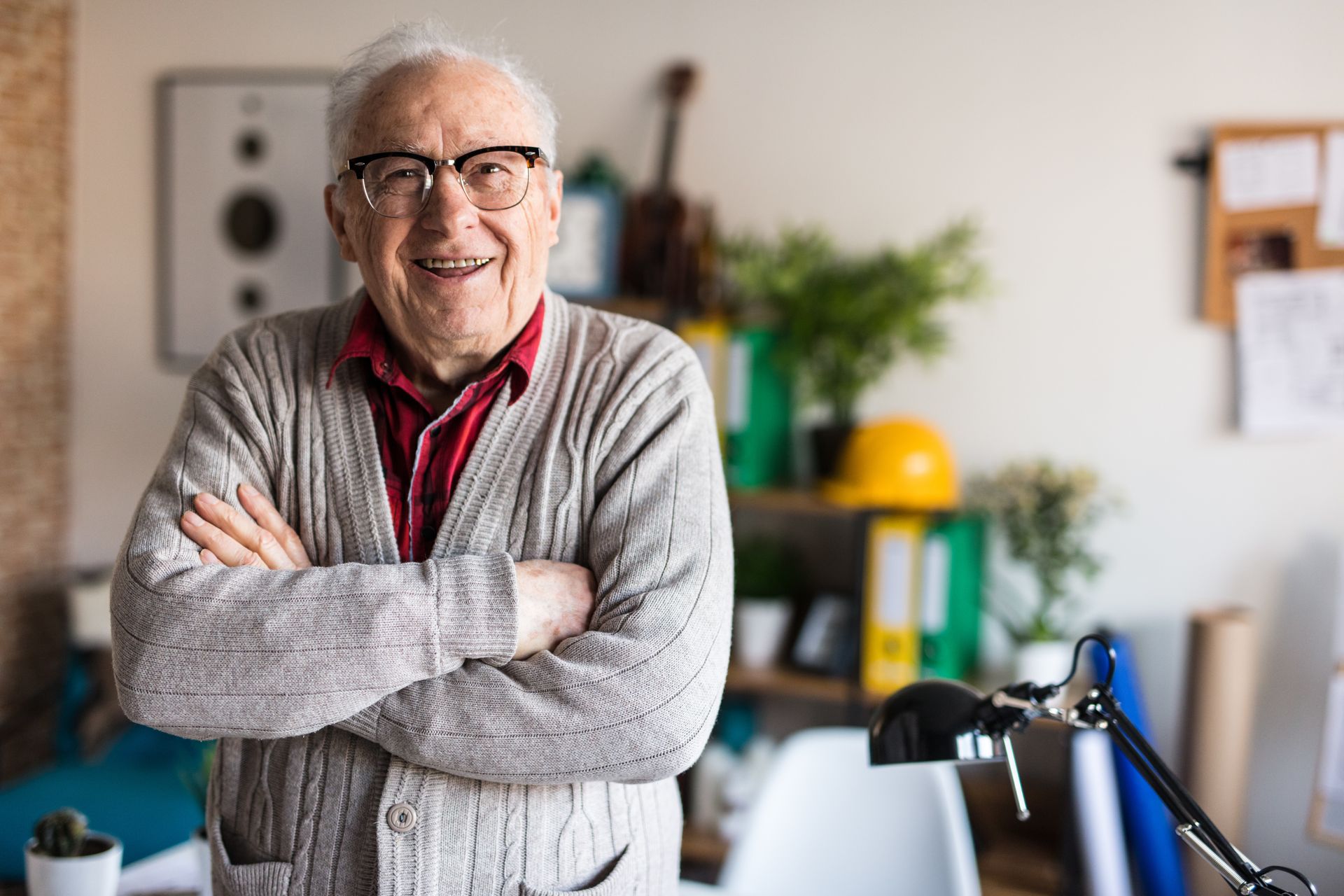 Older person in glasses, smiling, arms crossed. Standing in office space.