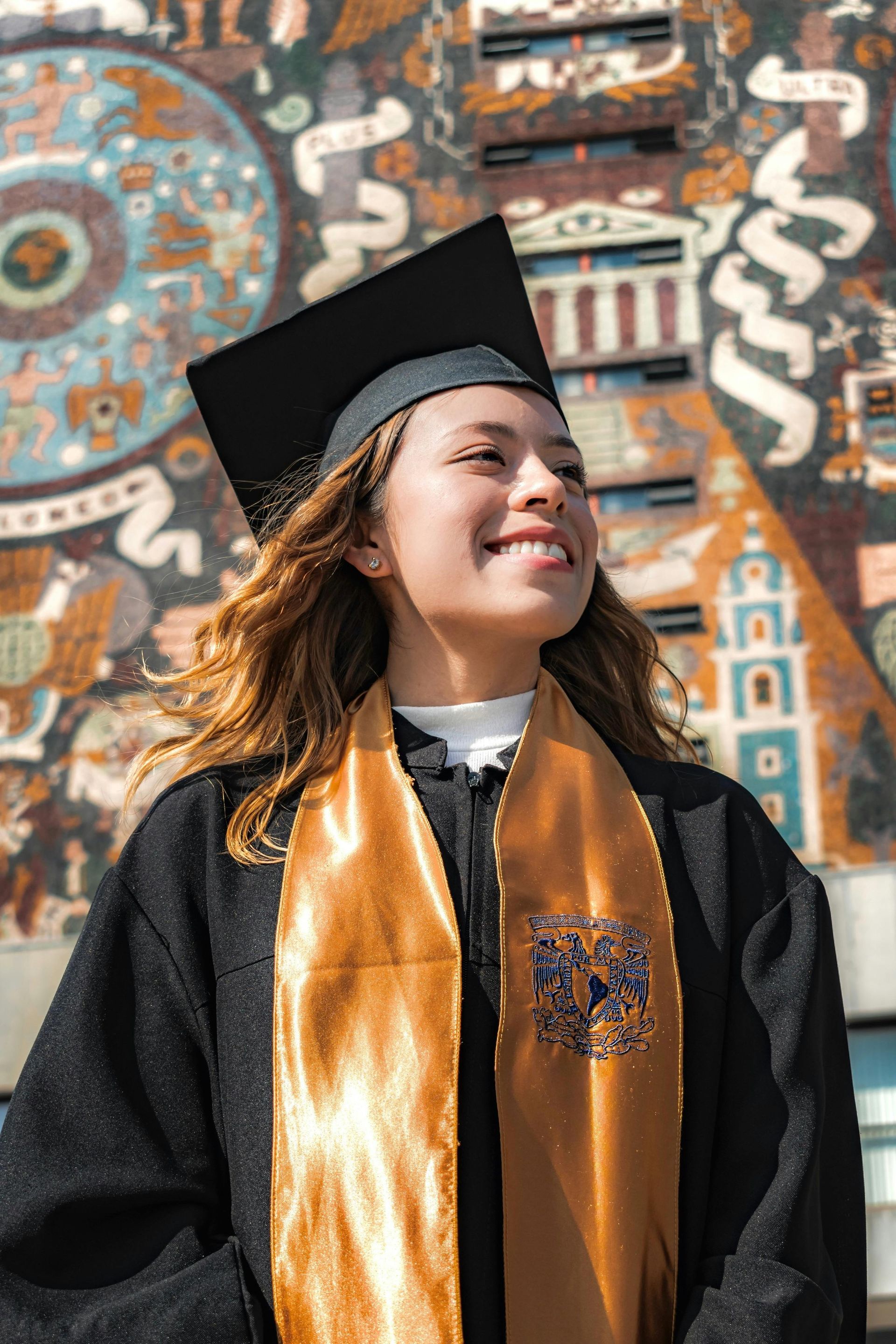 Graduating student in cap and gown, smiling, with gold sash, in front of a colorful mural.
