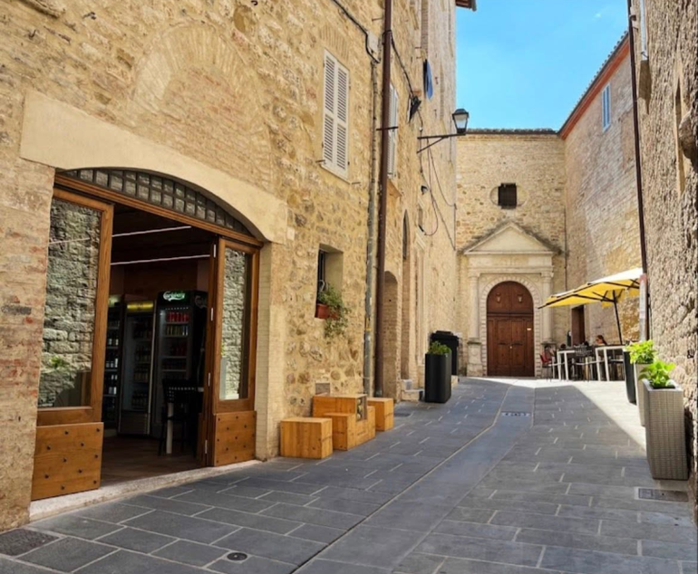 Un vicolo lastricato con edifici ad arco, tavolini da caffè e una piccola chiesa in fondo, sotto un cielo azzurro.