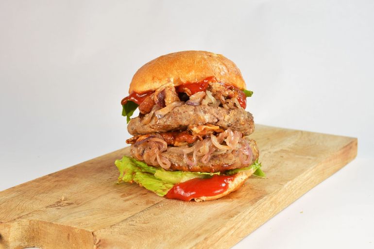 Hamburger impilato con lattuga e pomodoro su un tagliere di legno su sfondo bianco.