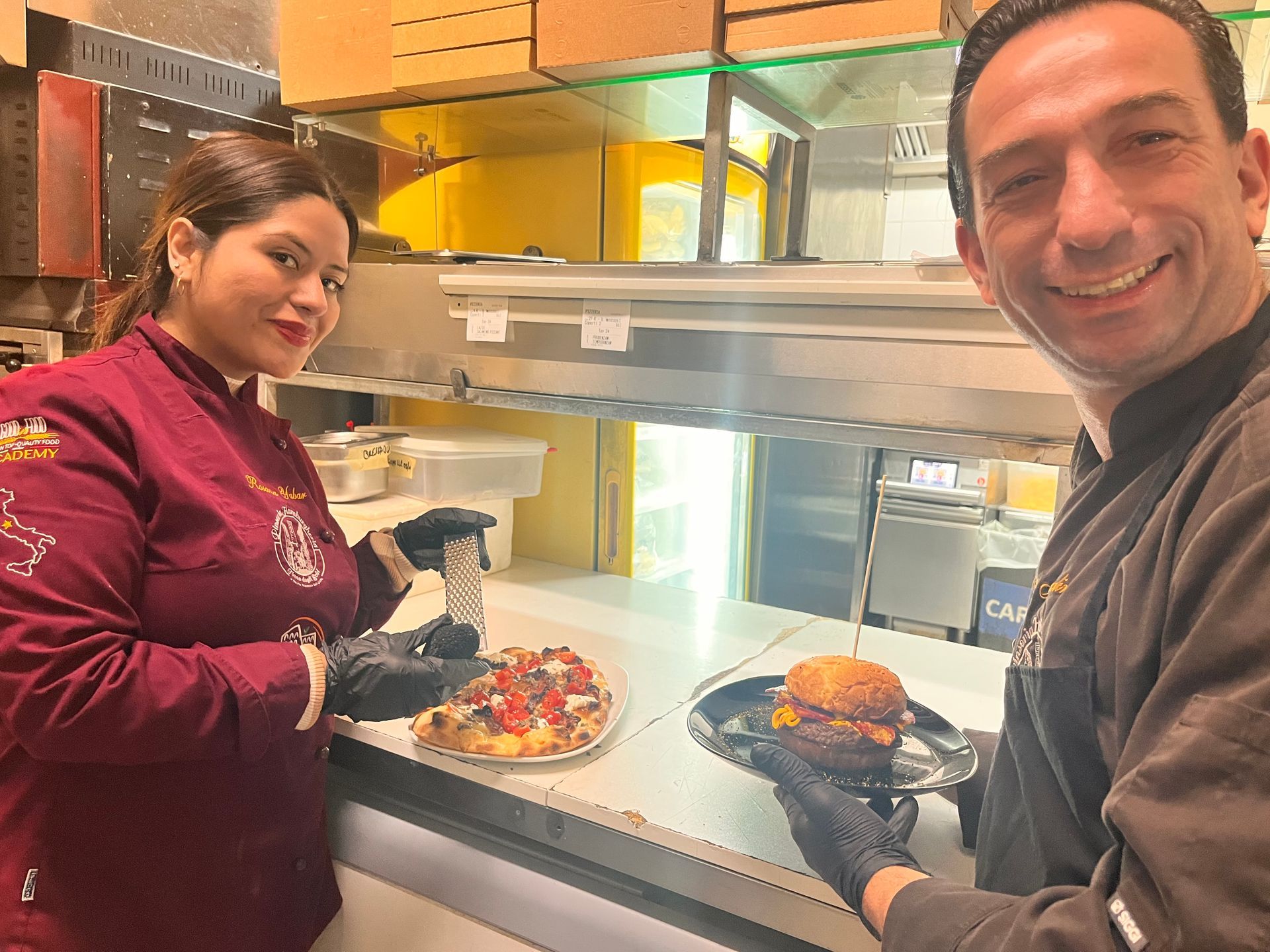 Due persone in cucina che tengono in mano dei piatti e sorridono alla telecamera.