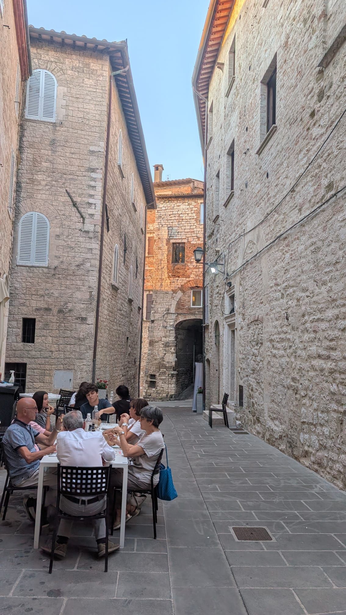 Persone che pranzano ai tavoli all'aperto in uno stretto vicolo di pietra tra alti edifici di mattoni.