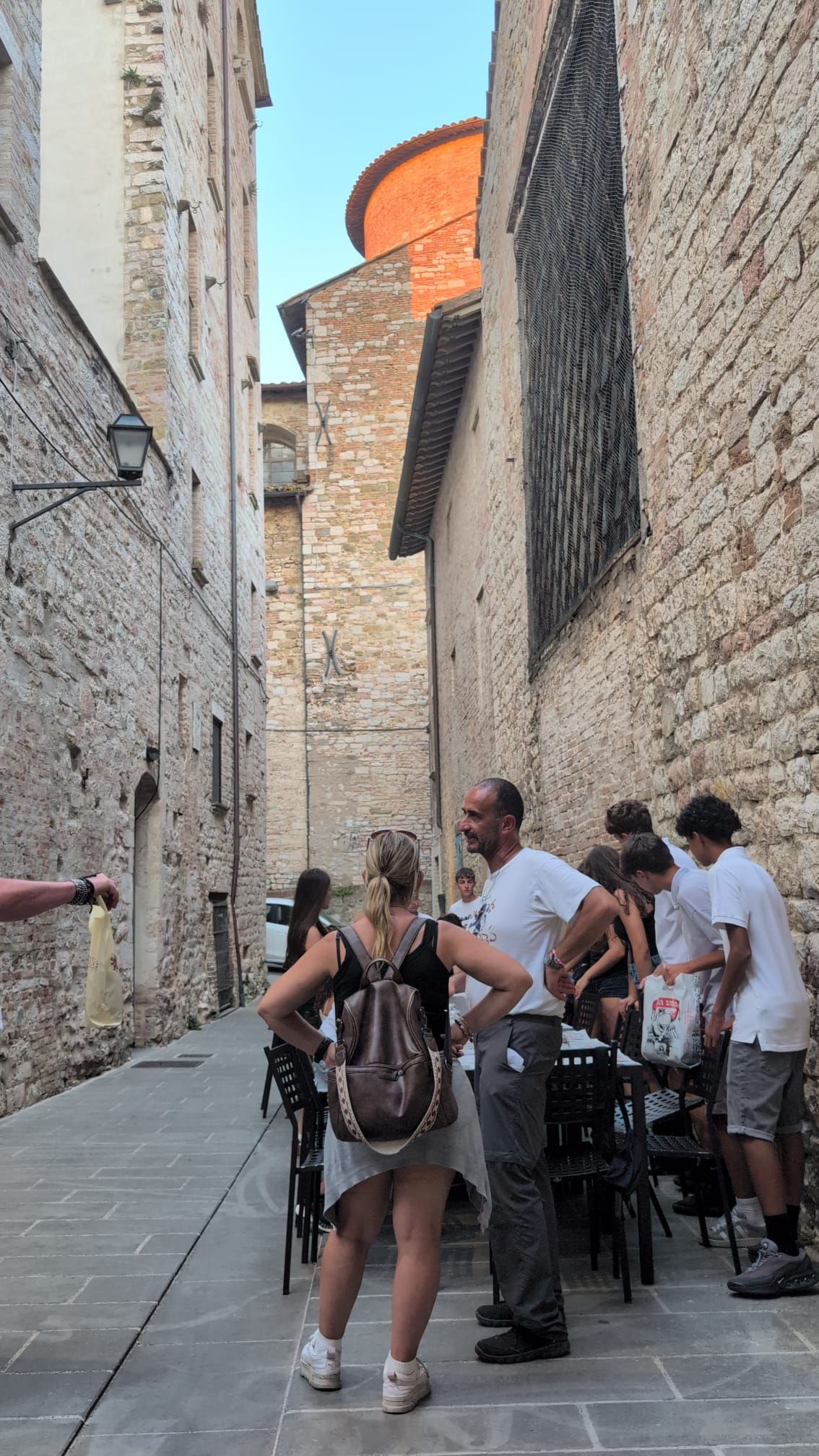 I turisti si sono radunati in uno stretto vicolo di pietra accanto a una torre storica e si sono seduti ai tavolini dei caffè all'aperto.