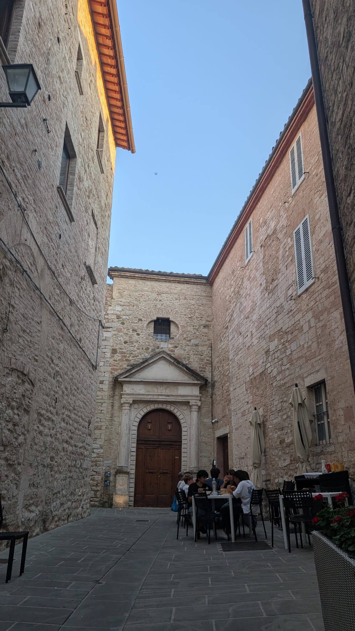 Un cortile in pietra tra alti edifici antichi, con un portone di legno ad arco e persone sedute ai tavolini del caffè.