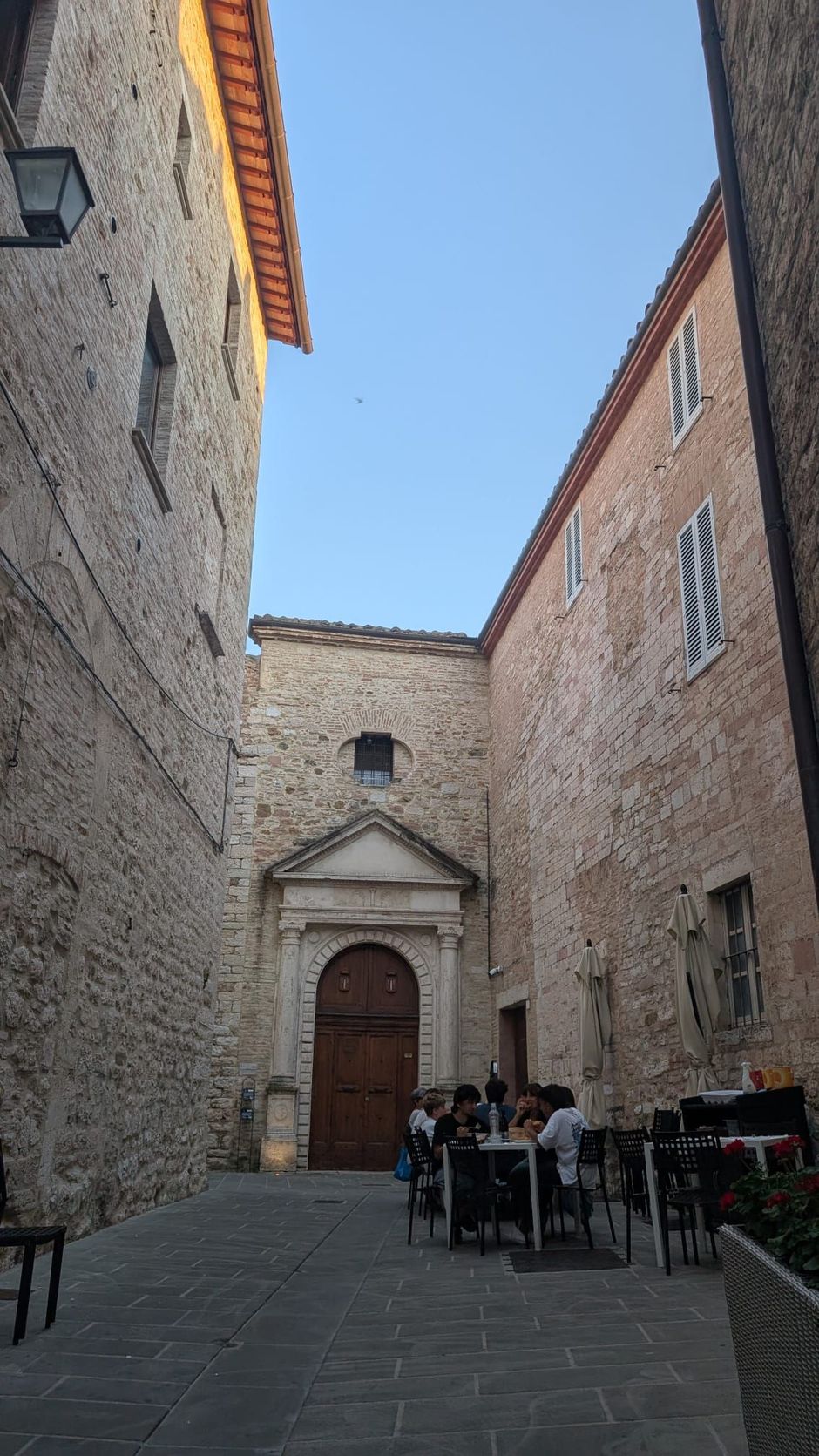 Stretto vicolo in pietra in un cortile storico con l'ingresso di una piccola chiesa e tavolini da caffè all'aperto.