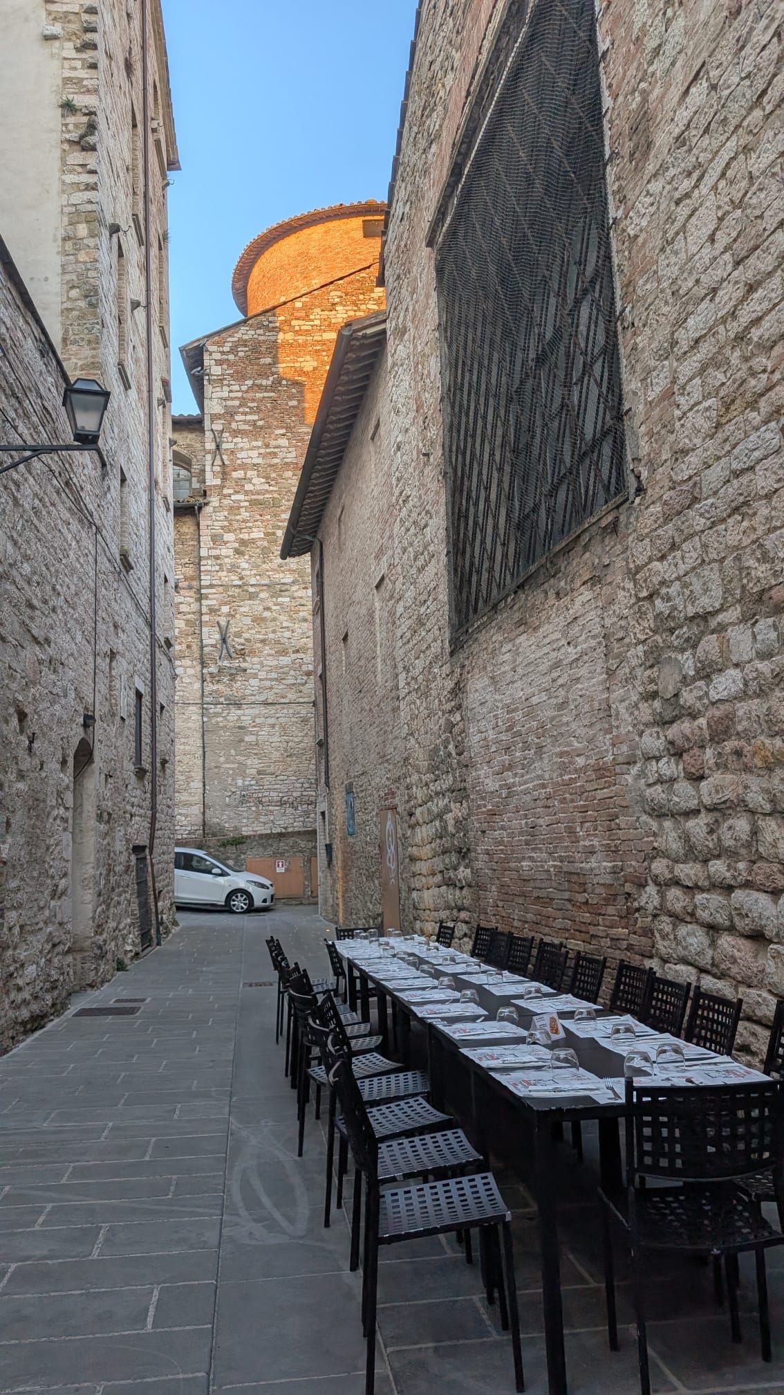 Stretto vicolo in pietra con tavolini e sedie da caffè all'aperto, accanto a un alto muro e a un'auto bianca parcheggiata.