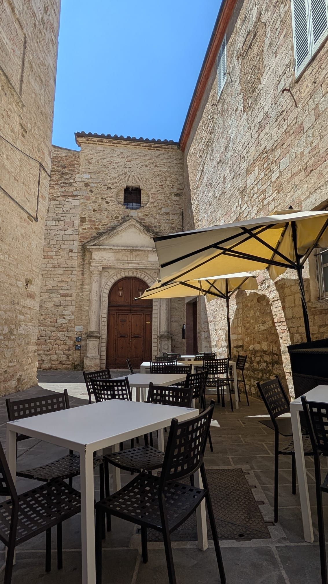 Caffè in un cortile di pietra con tavoli, sedie e grandi ombrelloni accanto a un'antica porta di legno, sotto un cielo azzurro.