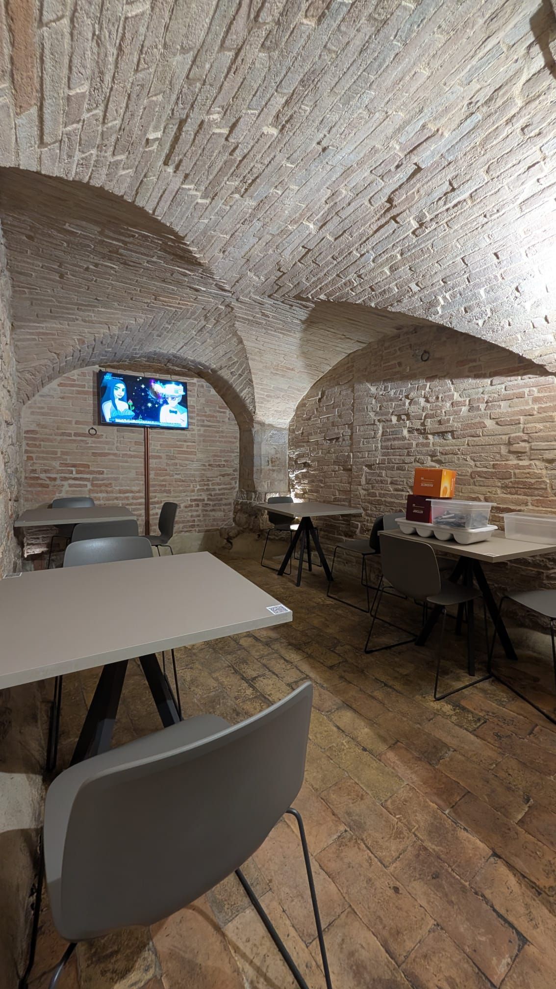 Sala con soffitto a volta in pietra, arredata con tavoli, sedie e un piccolo televisore montato a parete.