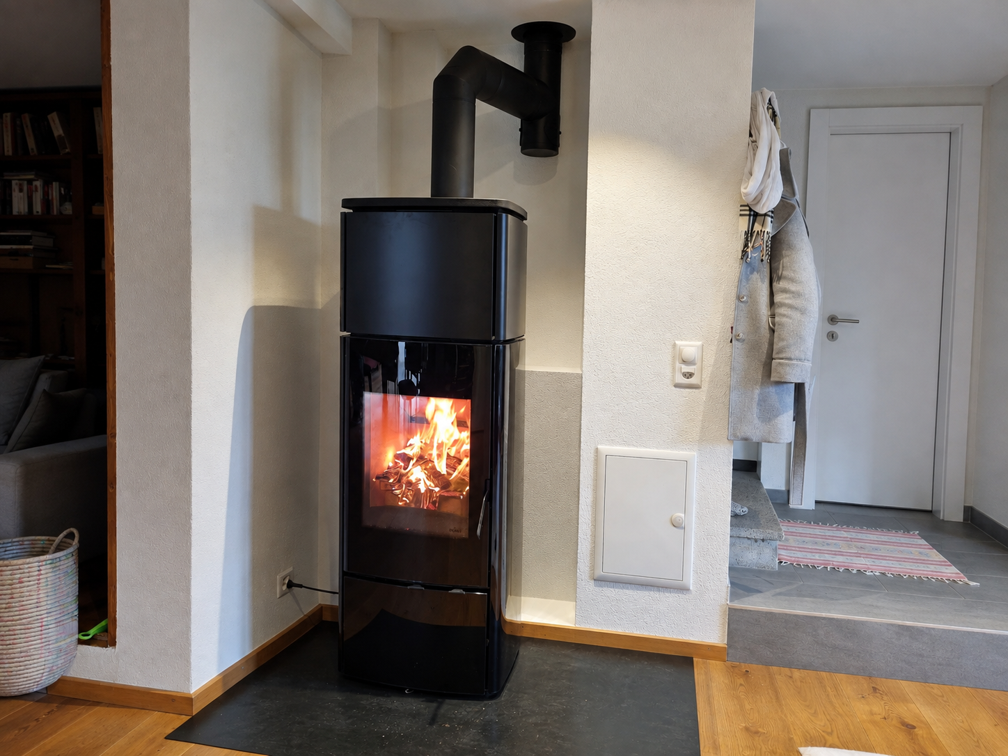 Poêle à bois noir avec un feu qui brûle dans un coin de la maison, près d'une porte et de manteaux suspendus.
