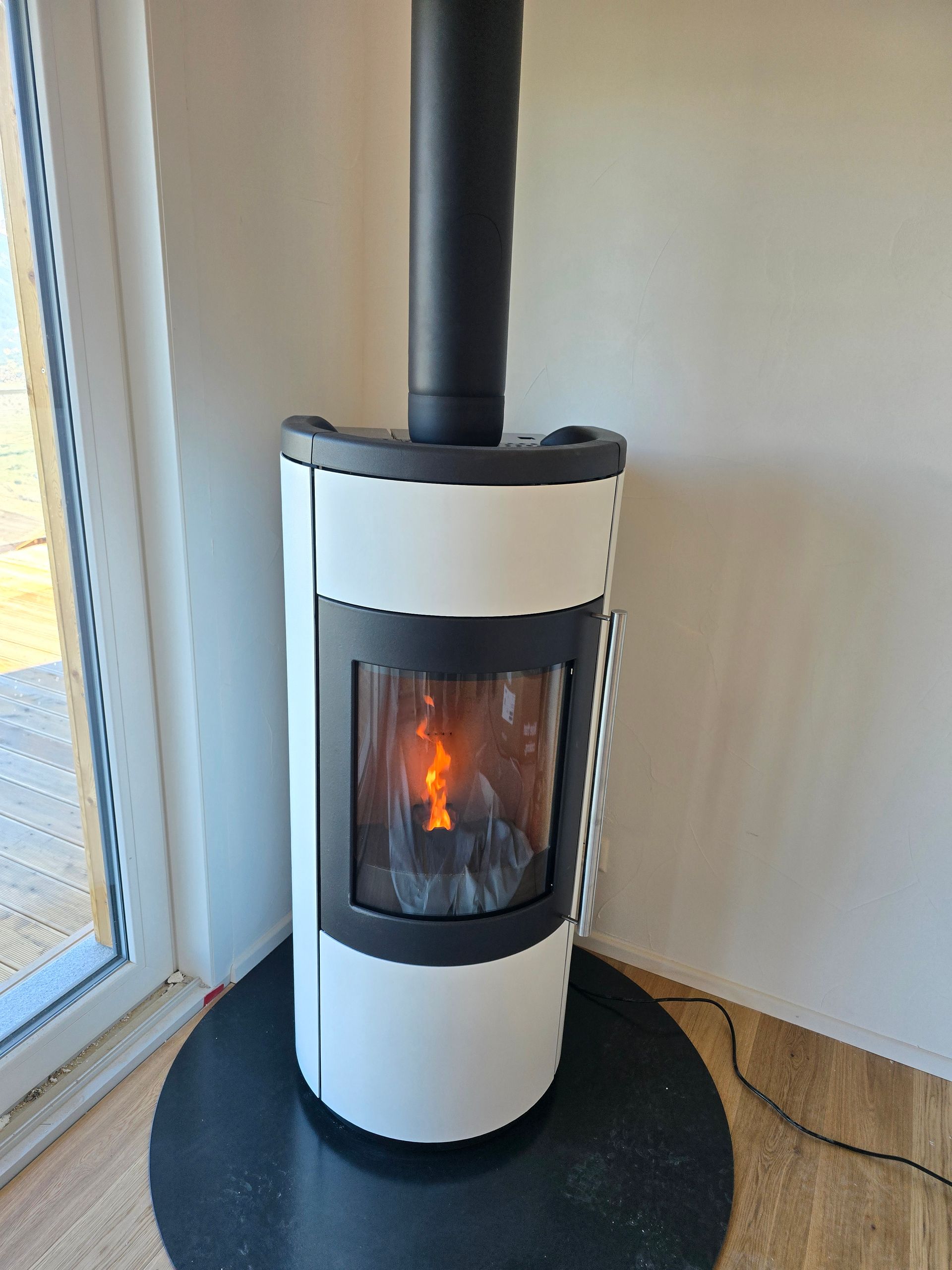Poêle à granulés cylindrique blanc avec un feu allumé, cheminée noire, sur un foyer rond noir près d'une fenêtre.