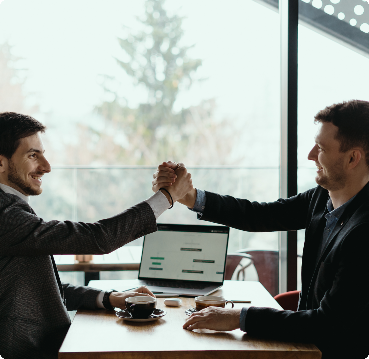 Two men are shaking hands while sitting at a table with a laptop.