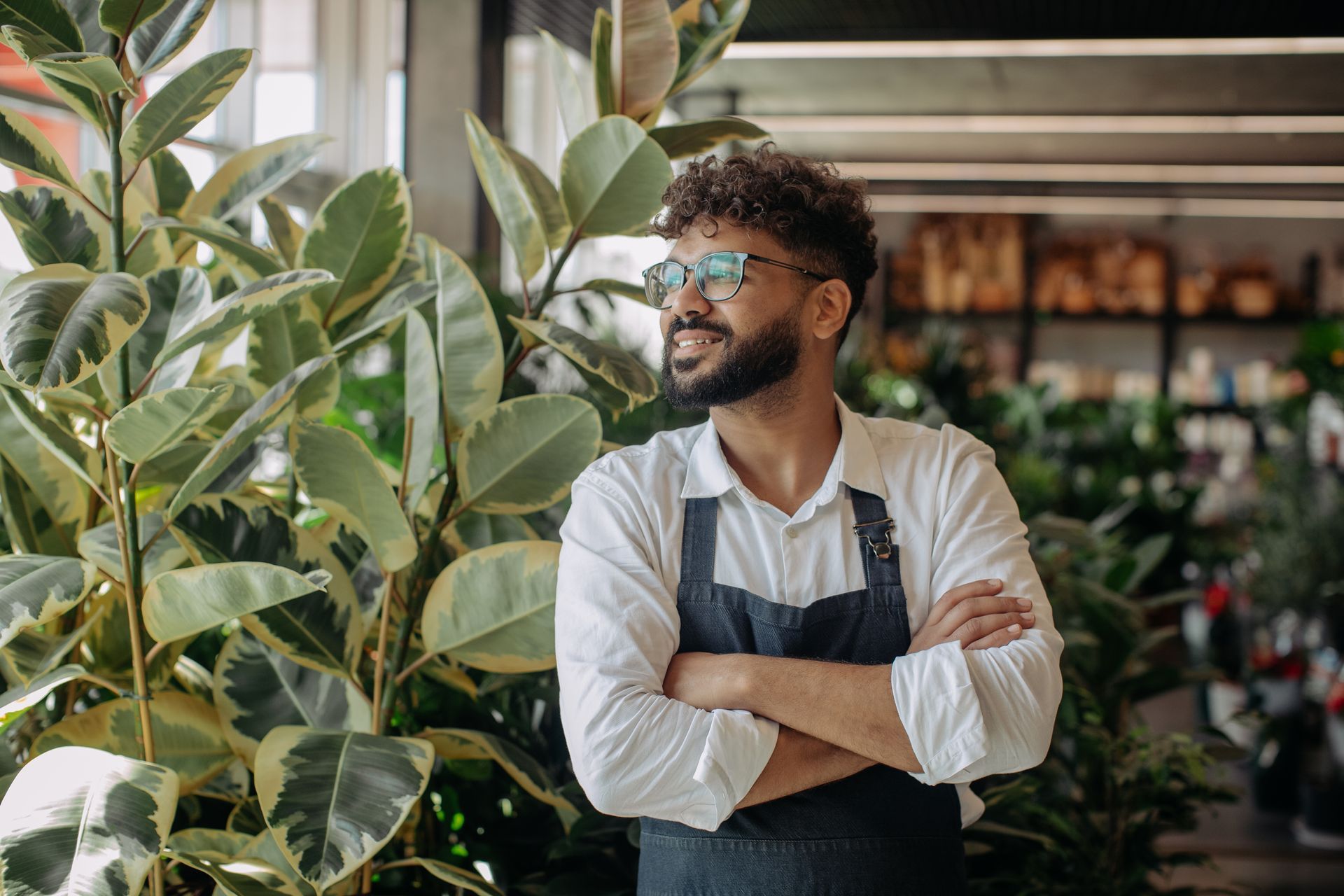 Man in apron and glasses stands in front of plants, arms crossed, in a shop.
