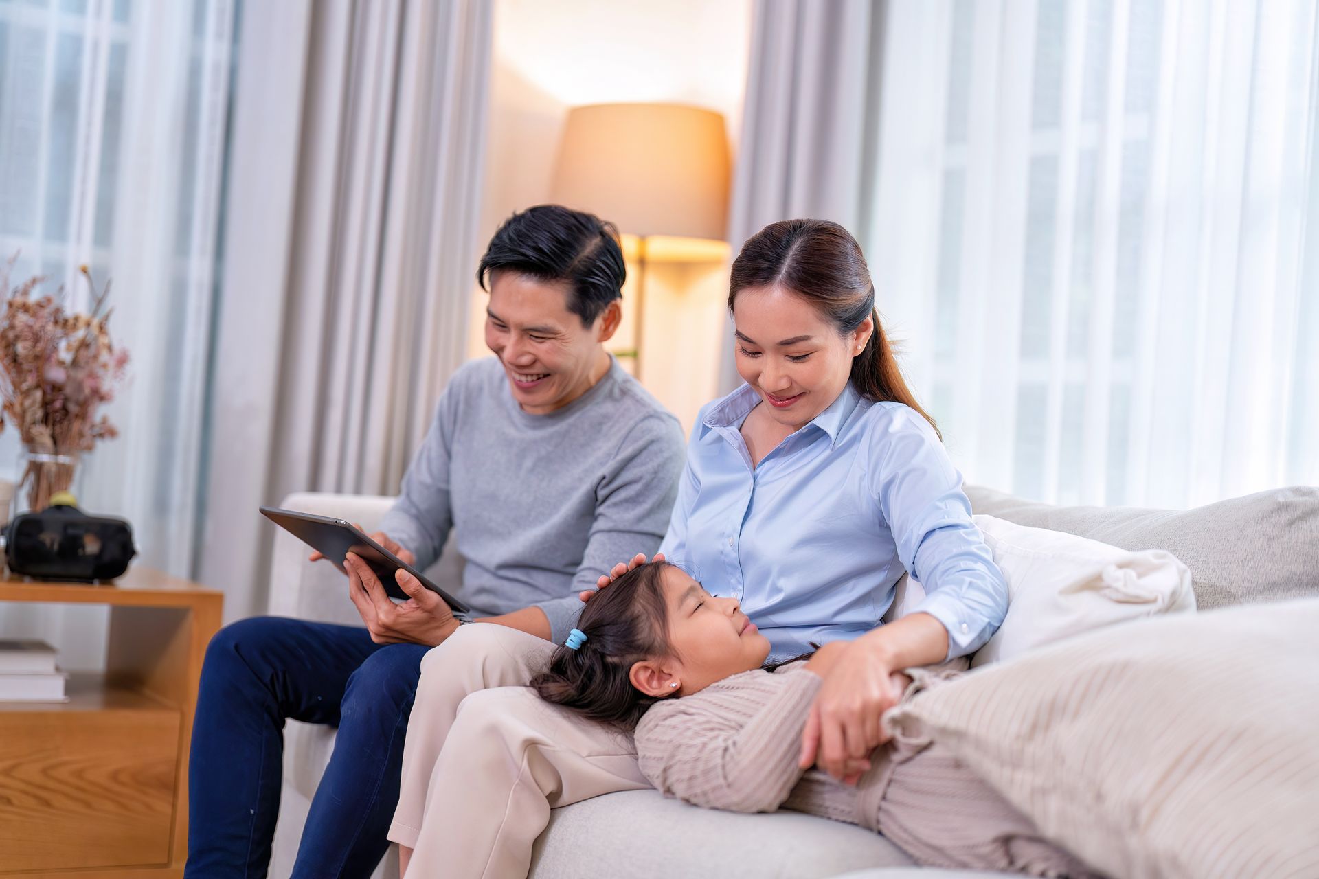 Family of three on a sofa, laughing; father reading tablet, mother stroking child's hair, lamp in background.