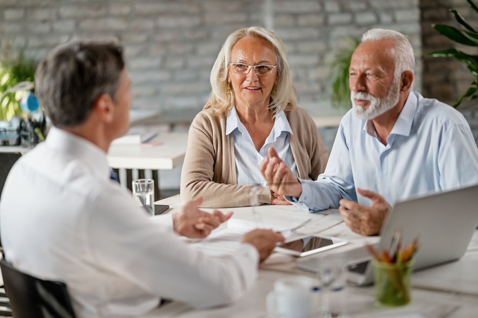 A financial advisor consults with an older couple at a table; documents and laptop present.