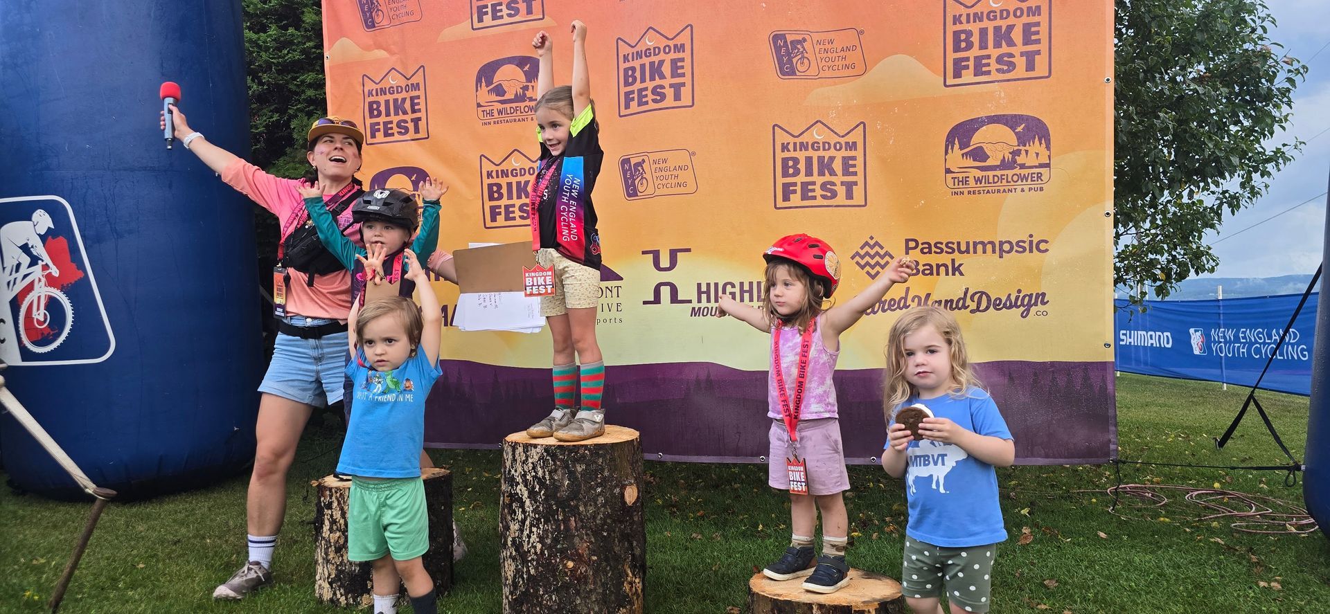 Children posing on a podium at an outdoor event, with medals, trophies, and a race backdrop.