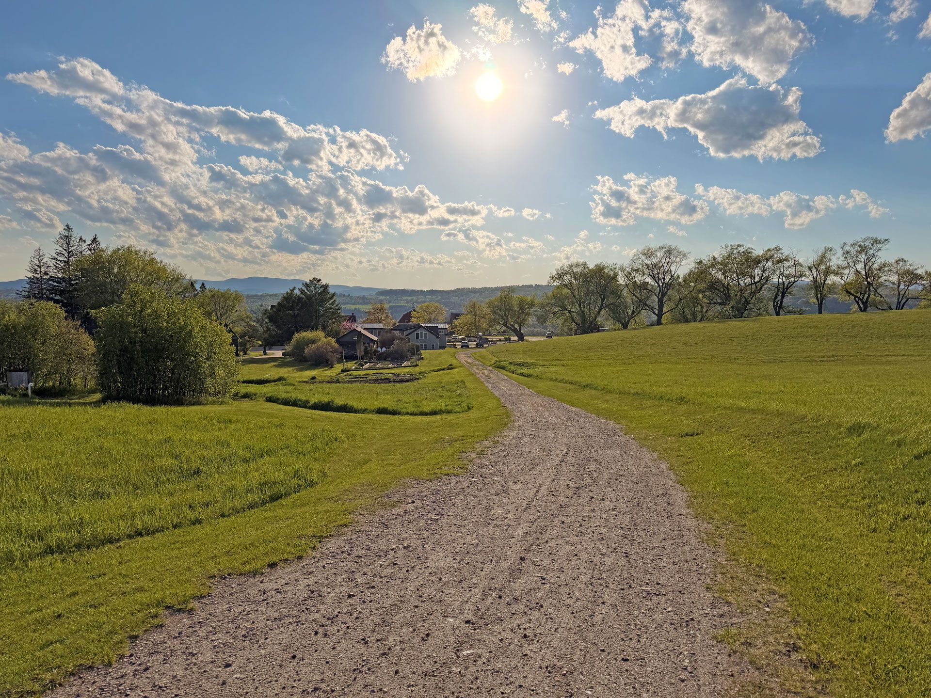 Sunlit gravel path through green fields toward a distant village under a bright, partly cloudy sky