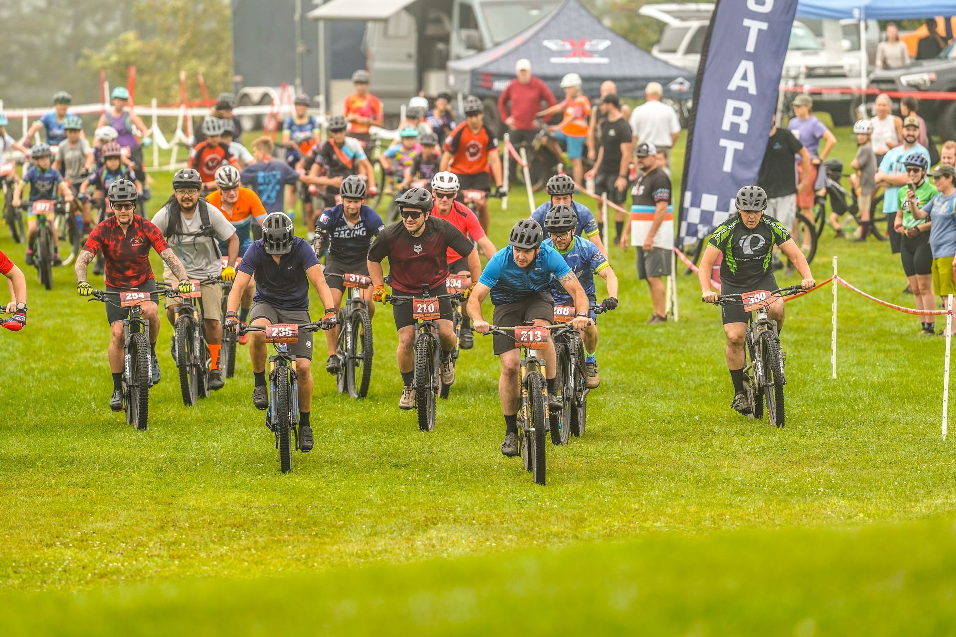 Mountain bikers racing on a grassy course, packed together at the start line in colorful gear.