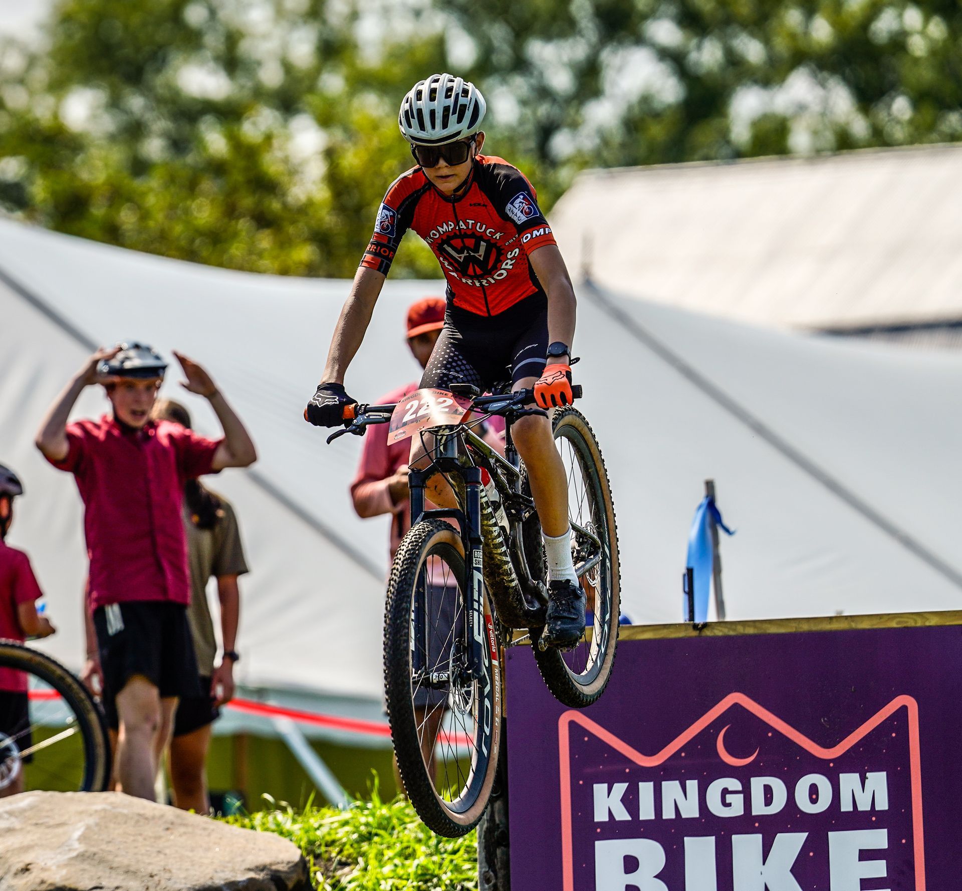 Mountain biker airborne over a purple ramp at a Kingdom Bike event, spectators watching nearby