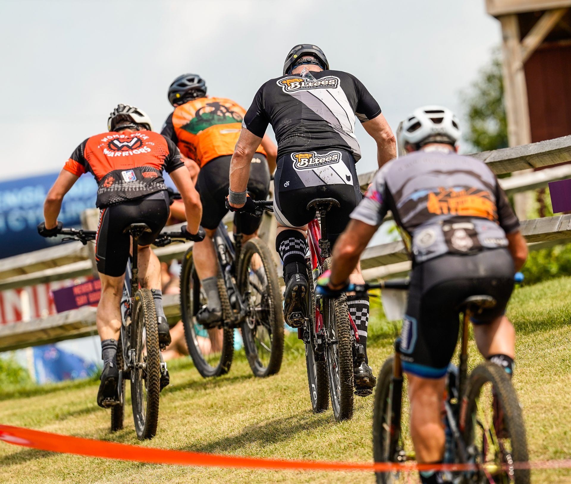 Cyclists racing uphill on a grassy course, seen from behind, with orange barriers and spectators in the background