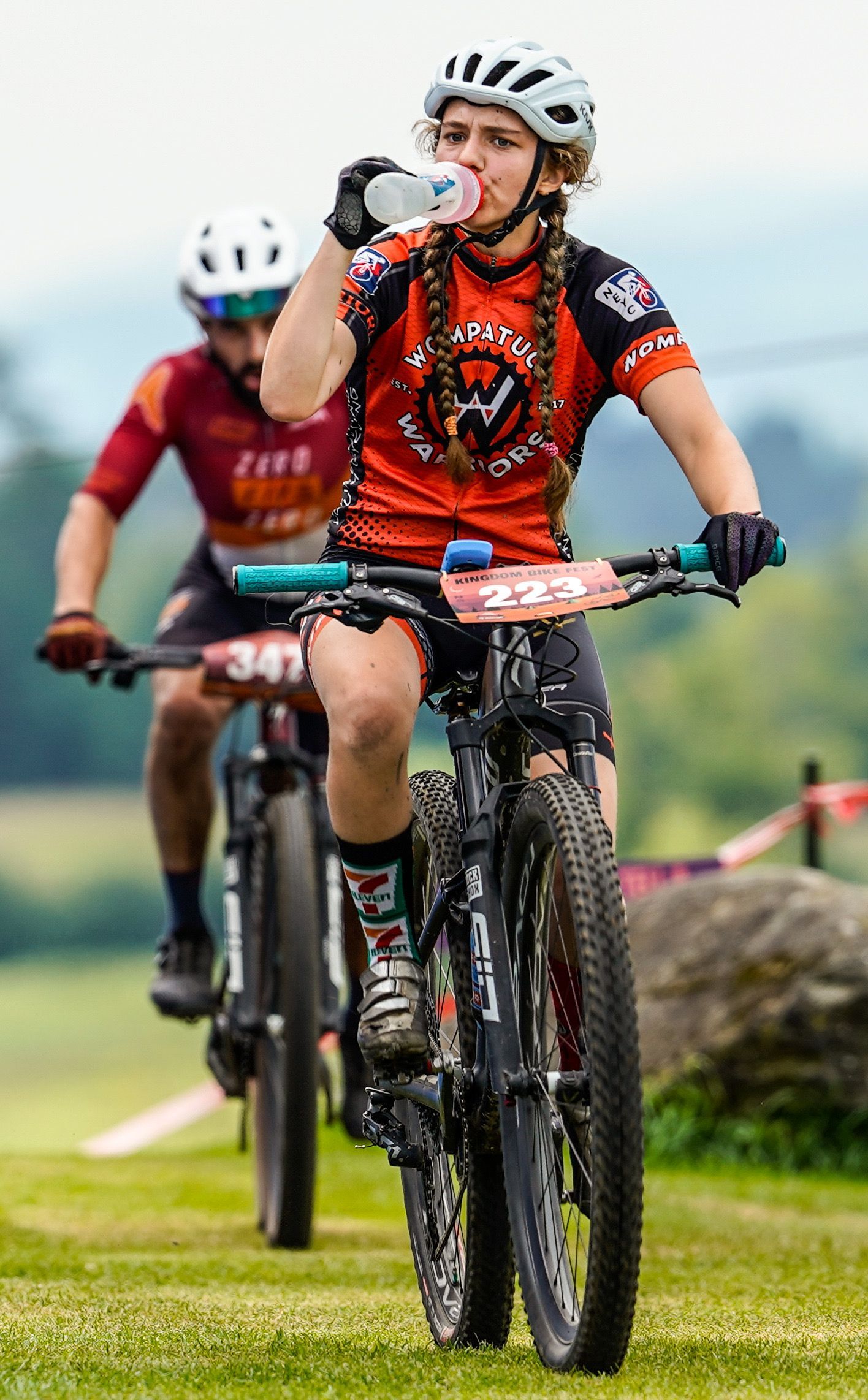 Two mountain bikers ride on a grassy trail, front rider in a red jersey and helmet, drinking from a bottle.
