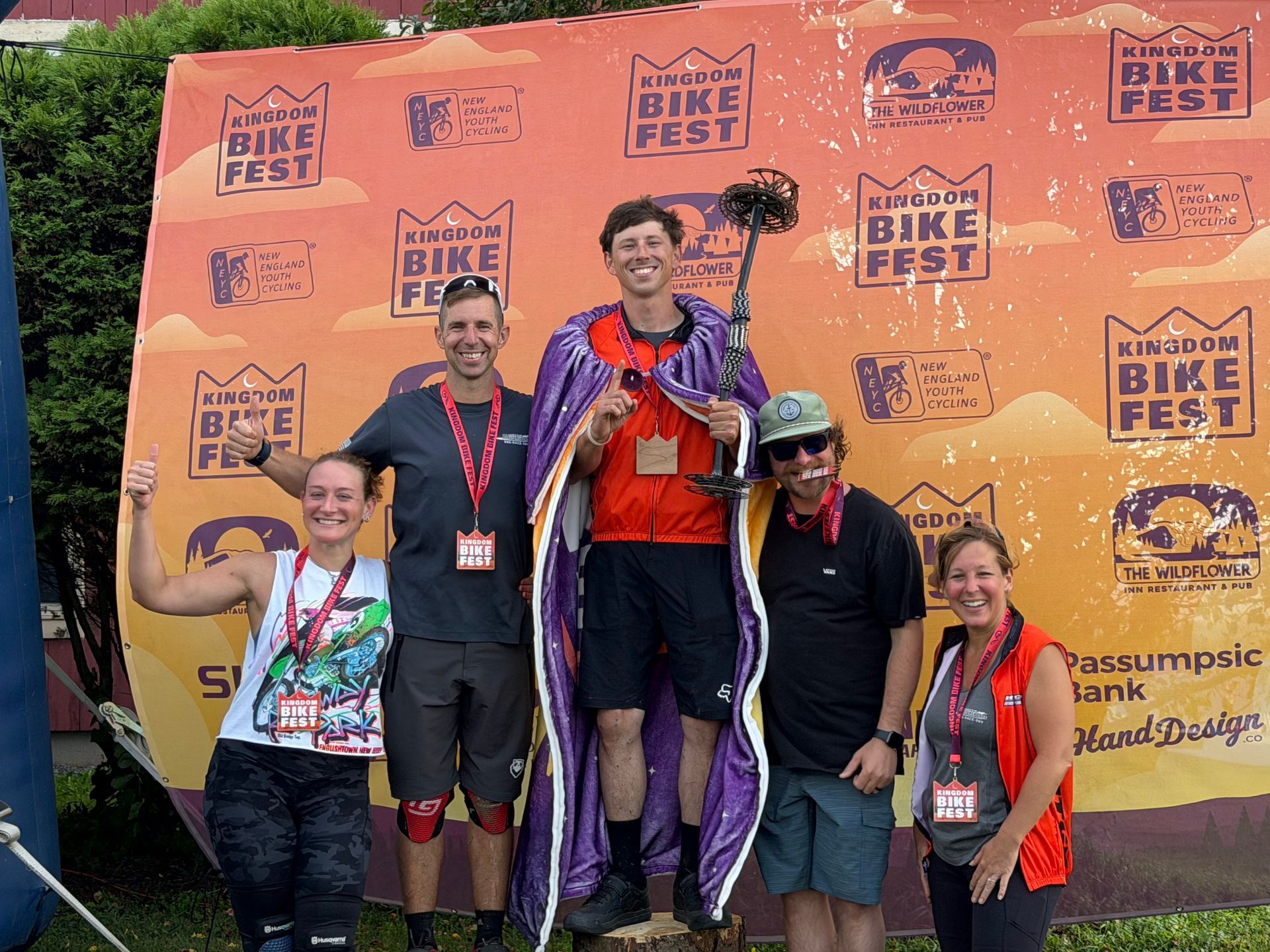 Five runners posing on a podium at Bike Fest, smiling with medals and trophies in front of an orange backdrop.