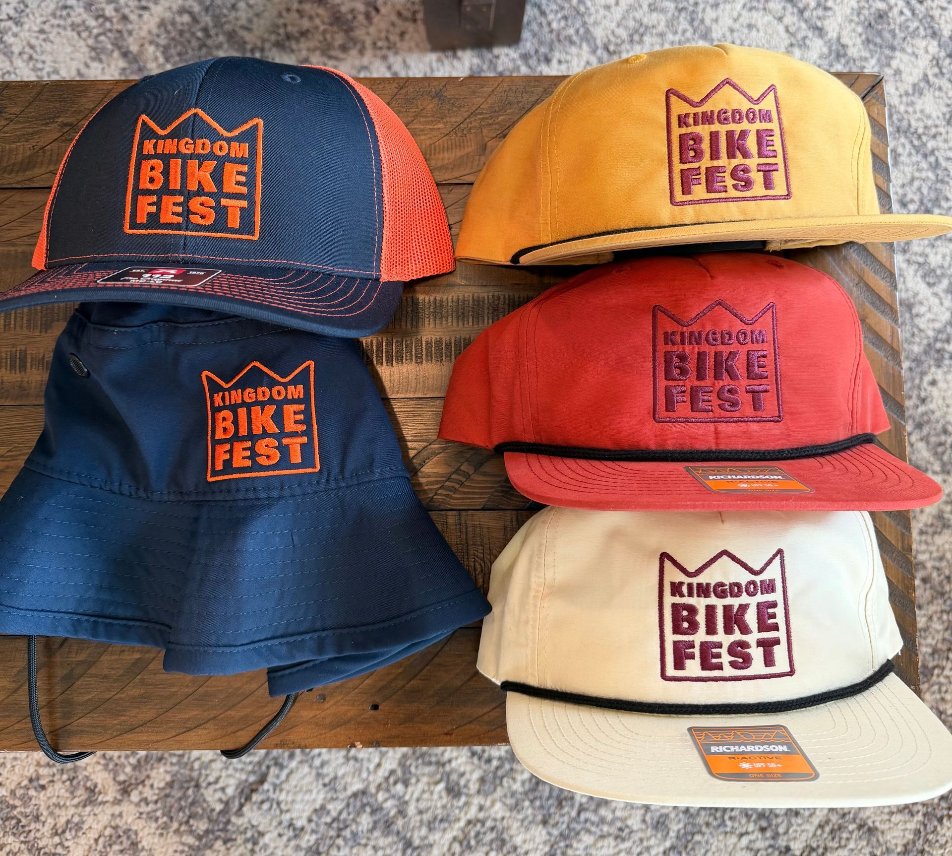 Five King of the Mountain Bike Fest hats in navy, yellow, red, and cream on a wooden surface