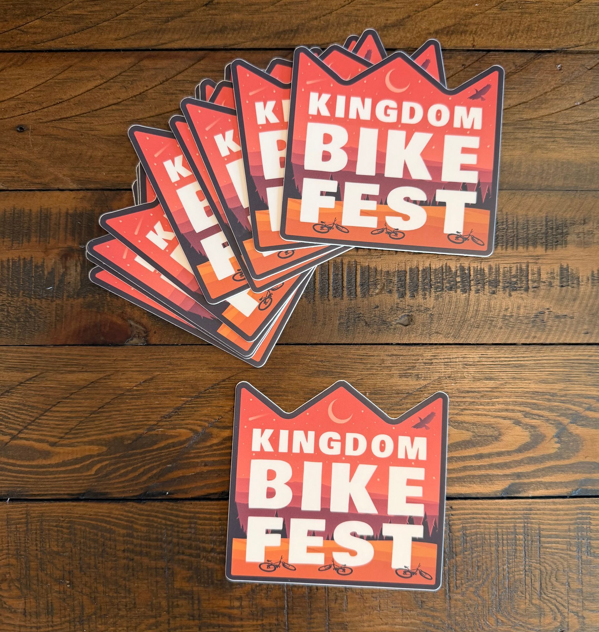 Stack of orange-and-red “Kingdom Bike Fest” stickers on a wooden table, with one sticker below.