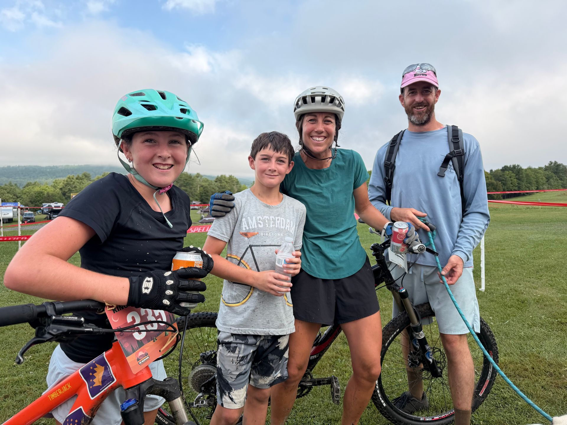 Four cyclists with bikes smiling outdoors on a grassy hill under a cloudy sky