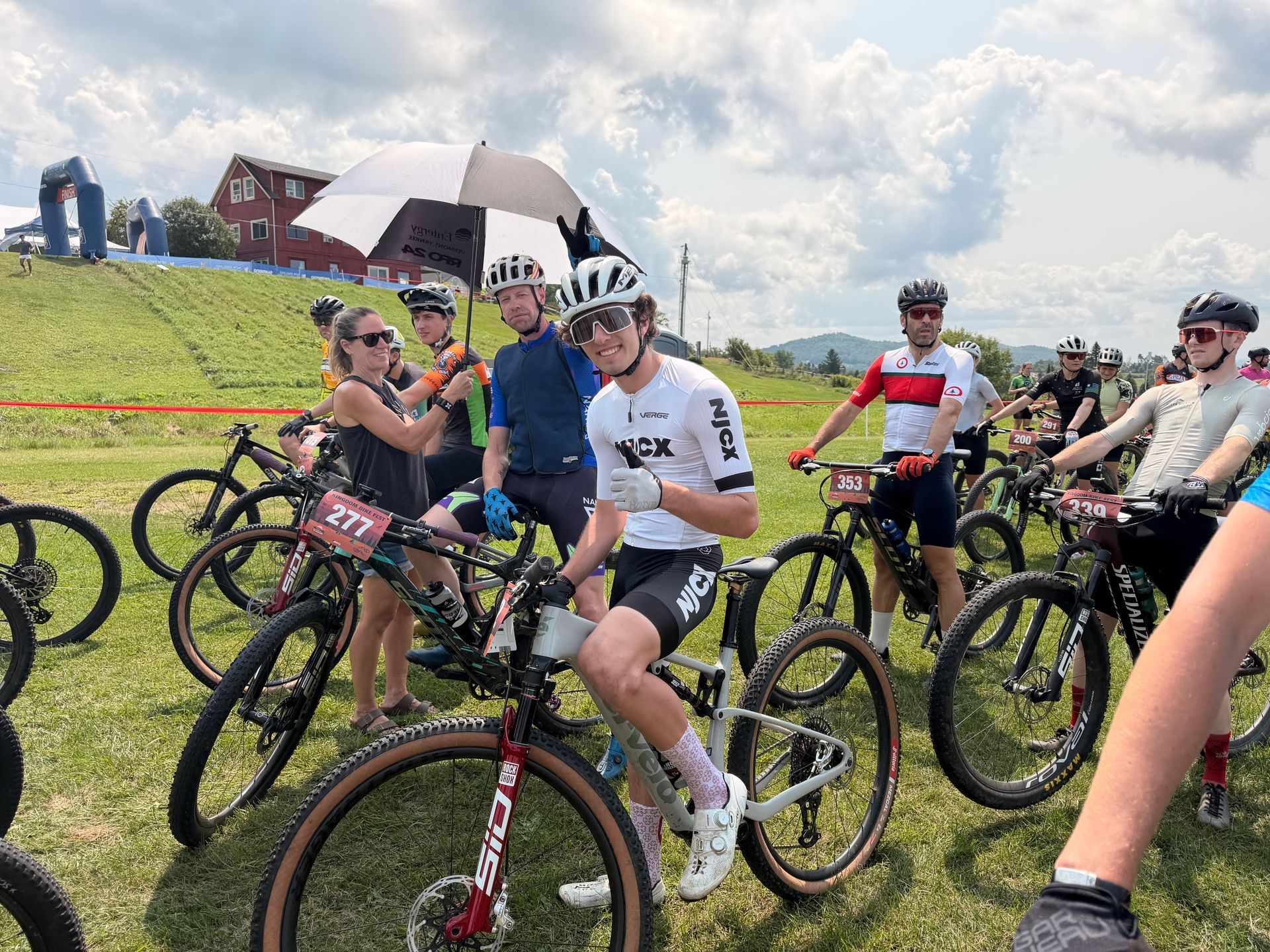 Cyclists gathered at a grassy start line with bikes and helmets under a cloudy sky.