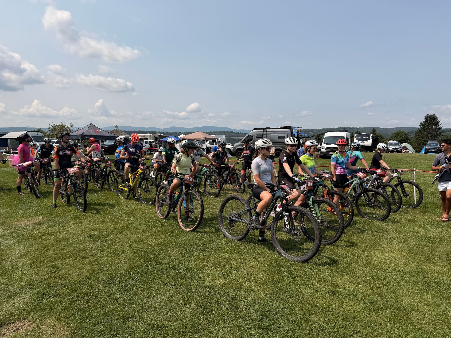 Cyclists gathered on a grassy field, with bikes lined up under a bright, cloudy sky.