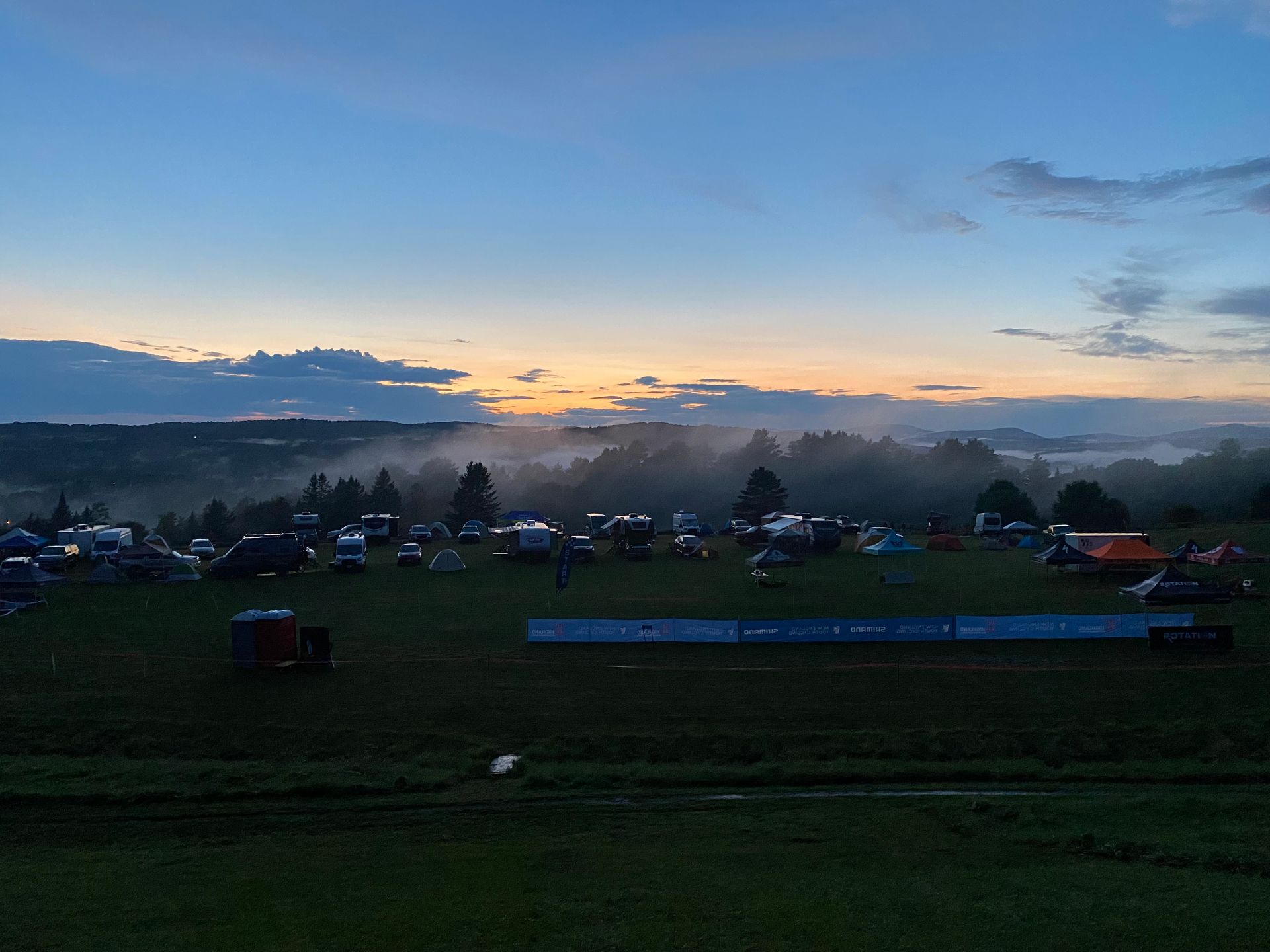 Dawn over a misty rural field with tents, parked cars, and distant hills under a blue-orange sky