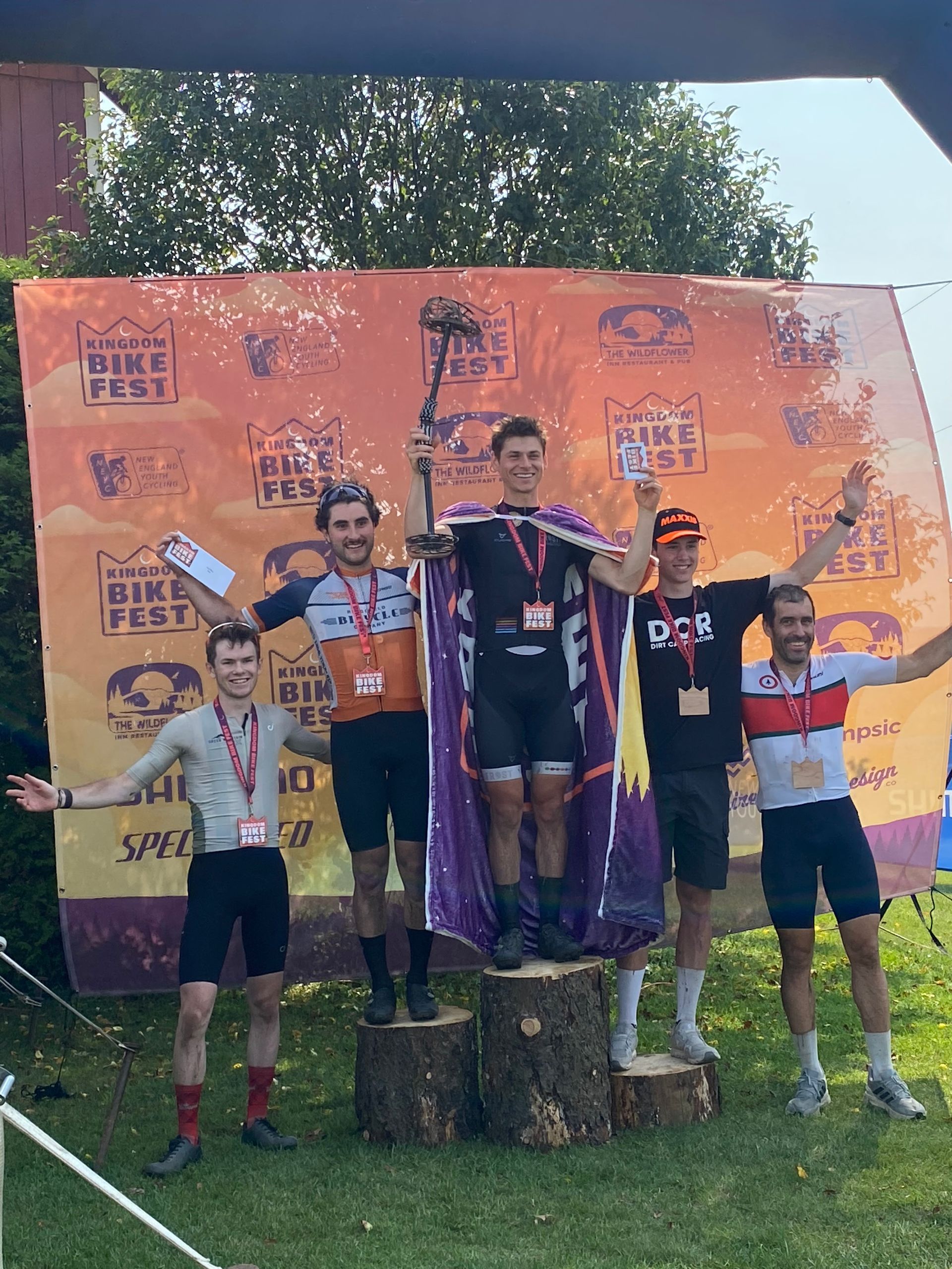 Five runners on a podium holding medals at an outdoor race awards ceremony.