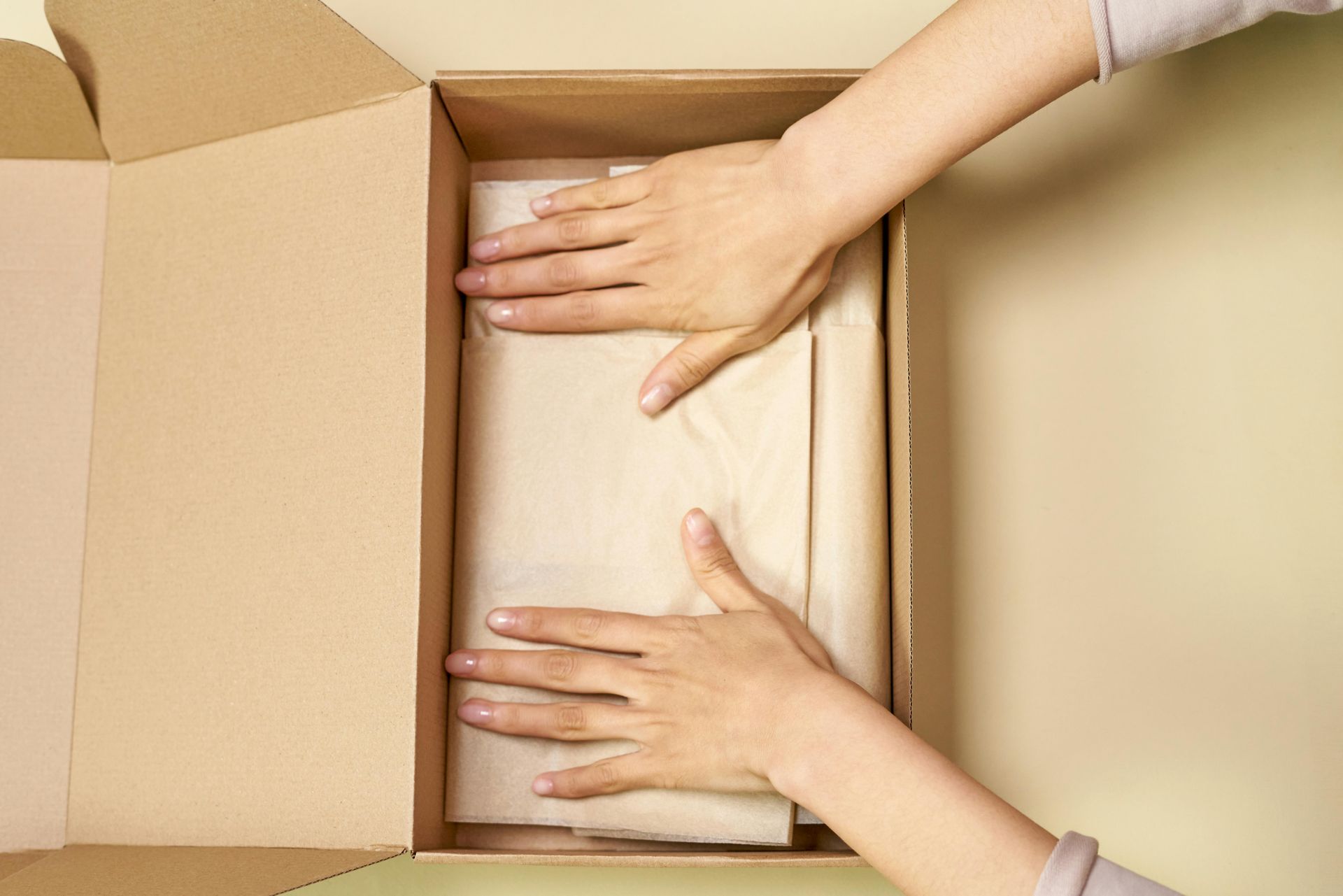 Handen pakken een ingepakt voorwerp in een kartonnen doos, lichtbeige achtergrond.