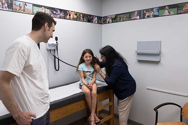 Child seated in exam room while clinician checks her, with another adult nearby