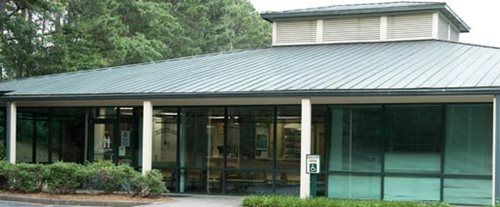Modern low building with glass windows and a green metal roof, surrounded by trees.