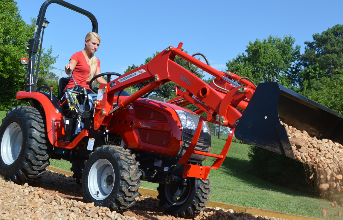 Woman operating a red tractor with a front-end loader carrying soil on a sunny day.
