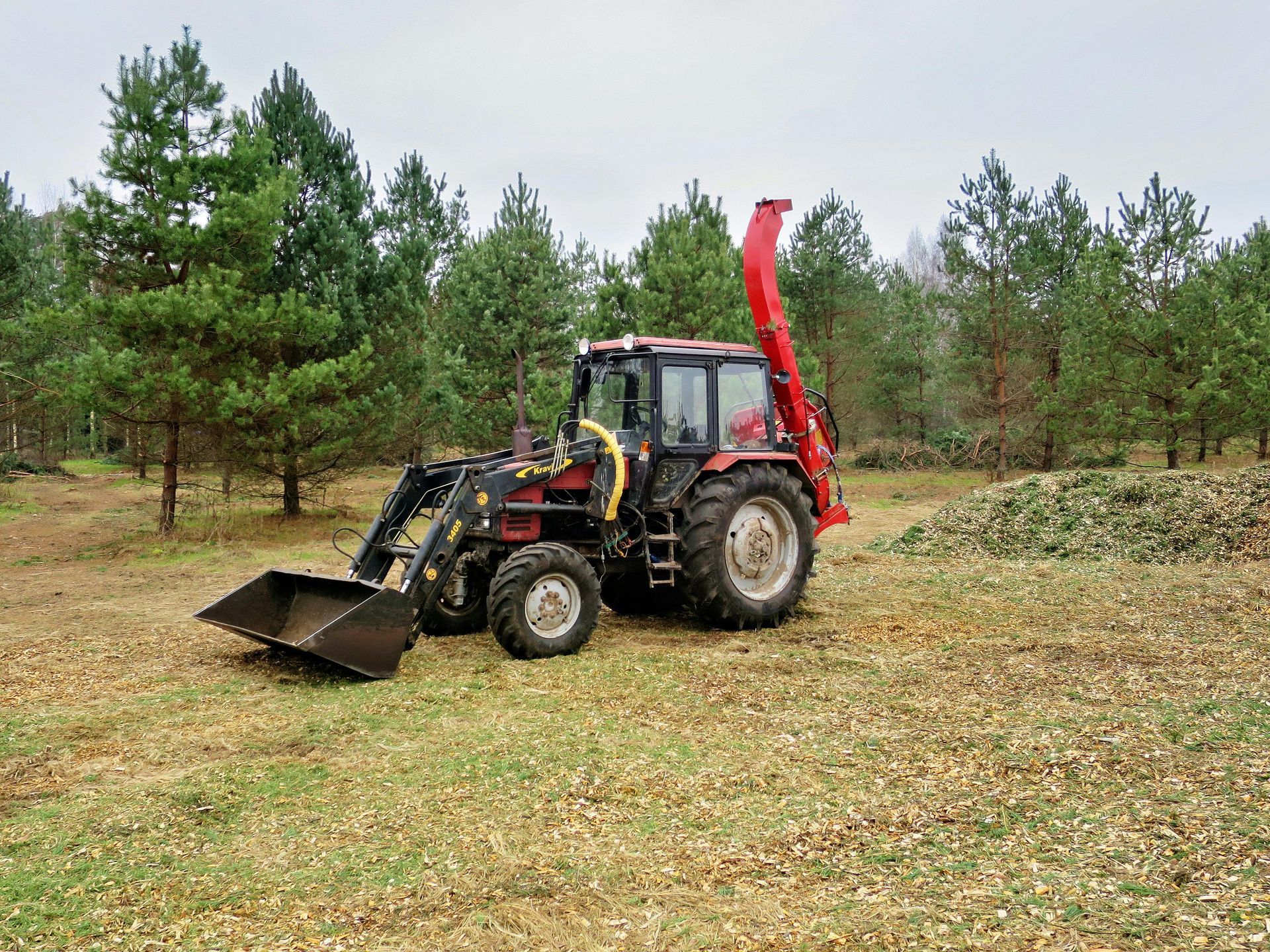 Tractor with front loader and red backhoe working in a grassy field with pine trees