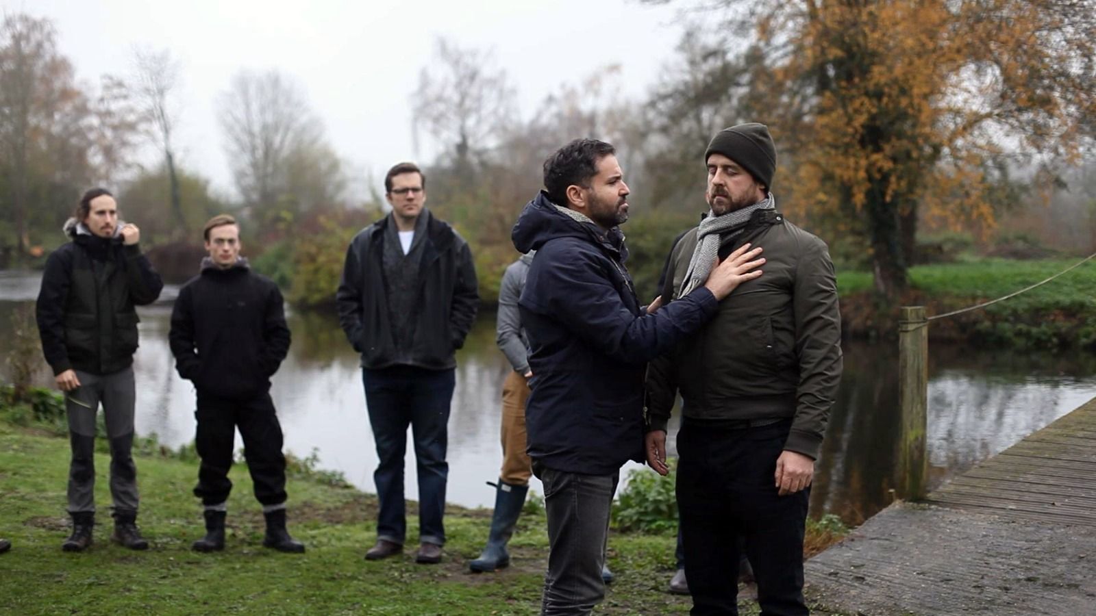 A group of people standing by a river in a wooded area, with one person gesturing toward another's chest.
