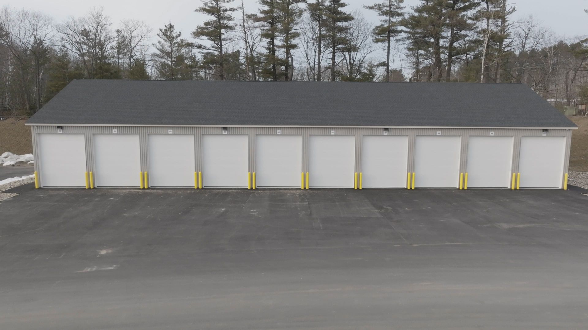 Row of storage units with white doors, gray roof, and asphalt parking area.