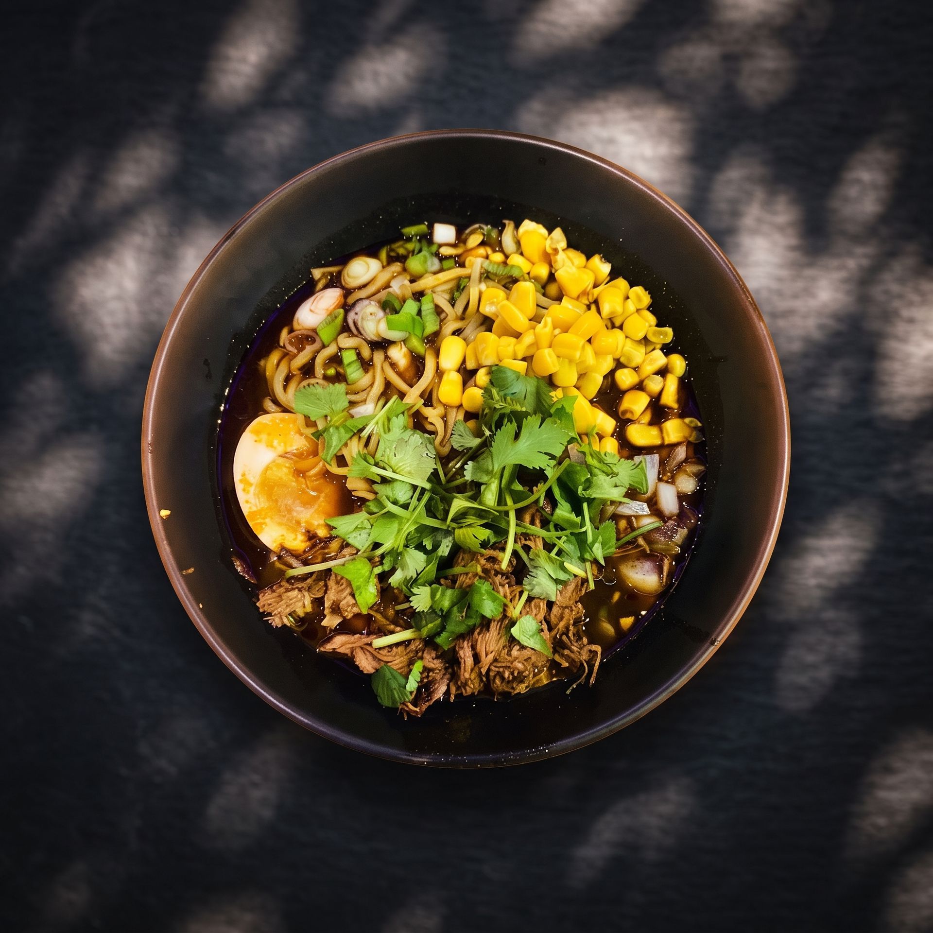 Black bowl of noodle soup topped with corn, herbs, and a soft-boiled egg on a dark table
