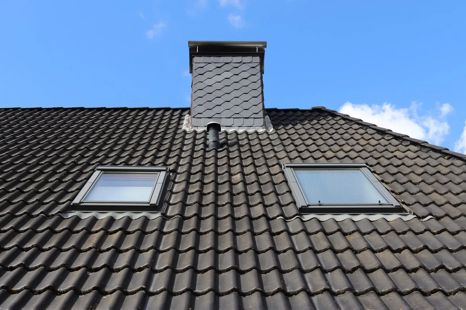 Dark tiled roof with chimney and two skylights against a blue sky.
