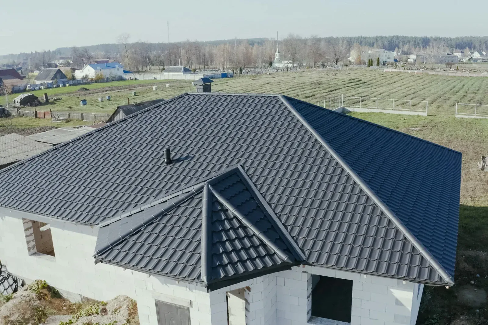 Gray-tiled roof of a house under construction; chimney and gabled roof section visible; rural setting.