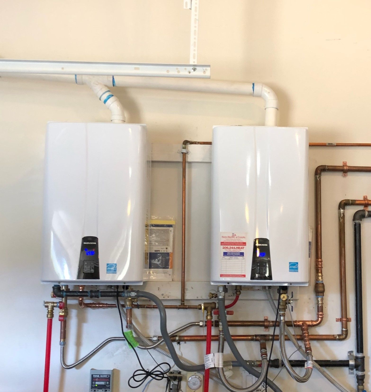Two white, wall-mounted tankless water heaters with copper piping against a light beige wall.