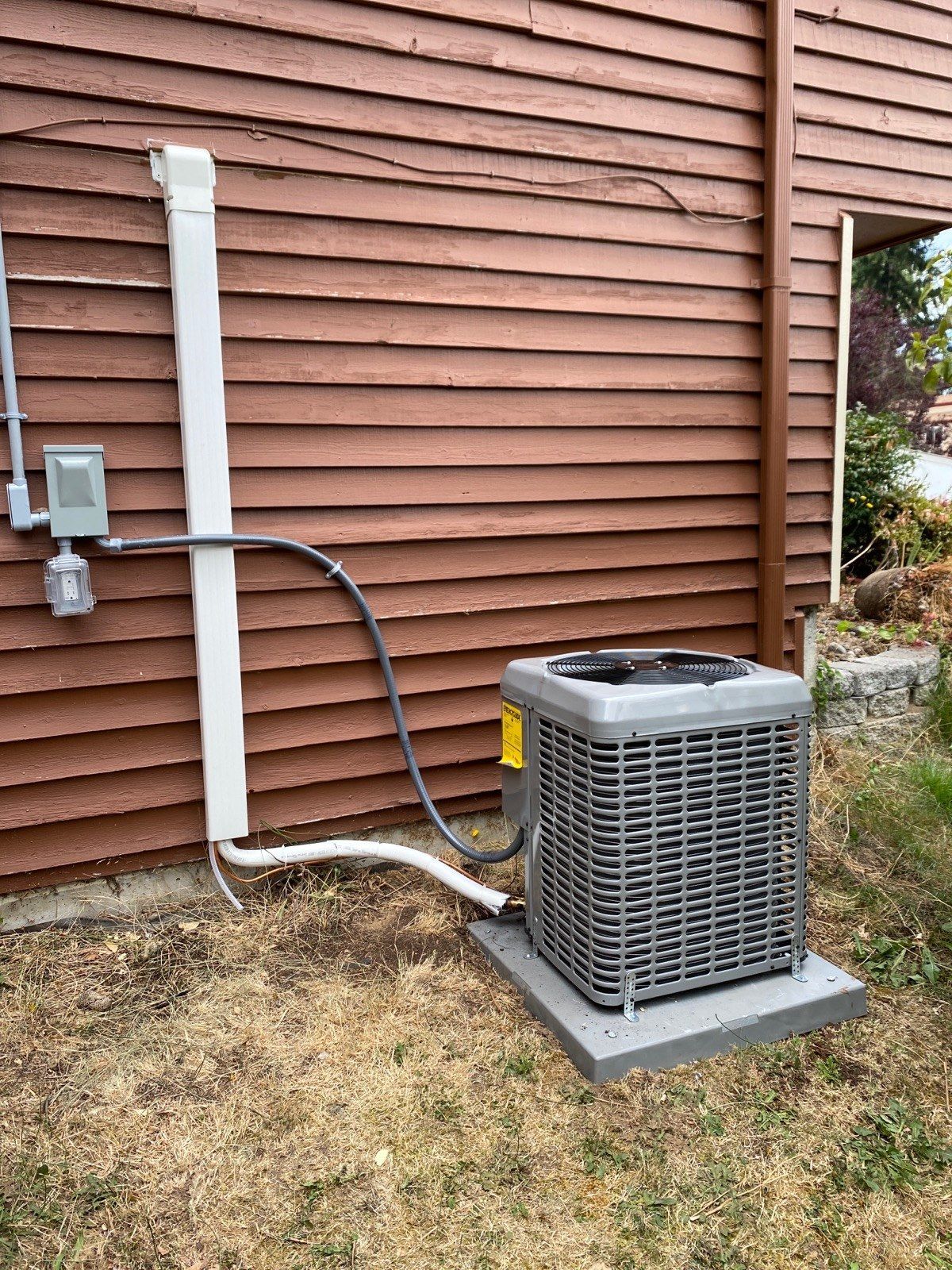 An air conditioning unit on a concrete pad outside a brown-sided building with electrical conduit.