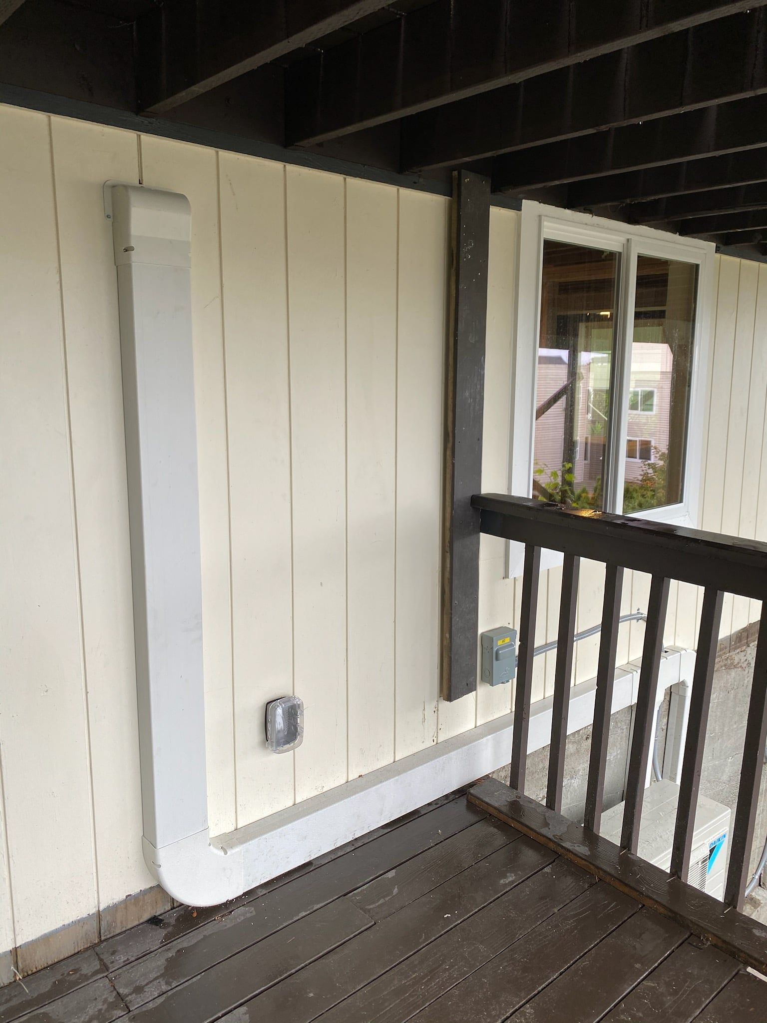 White conduit on a light beige wall of a deck, beside a window and railing.