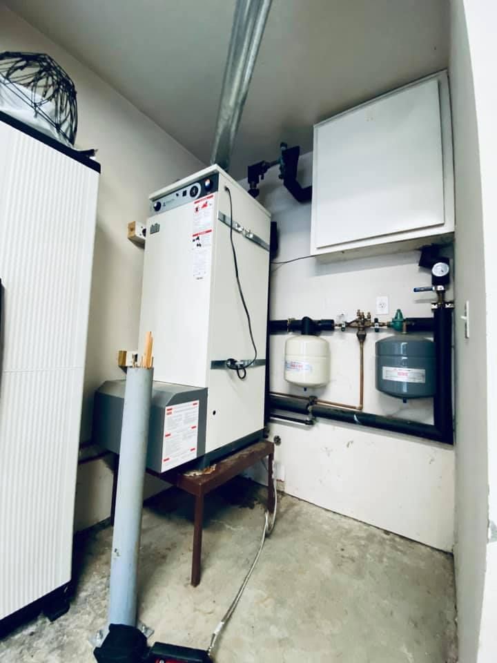 A boiler system in a utility room. White and gray machinery, pipes, and tanks mounted on walls.