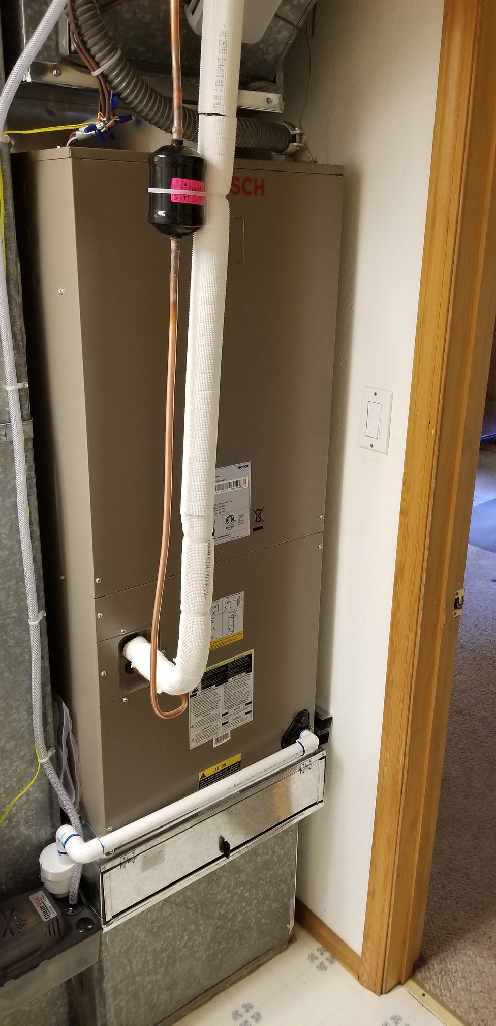 A beige furnace unit with white pipes and a black component in a room with a wooden door frame.