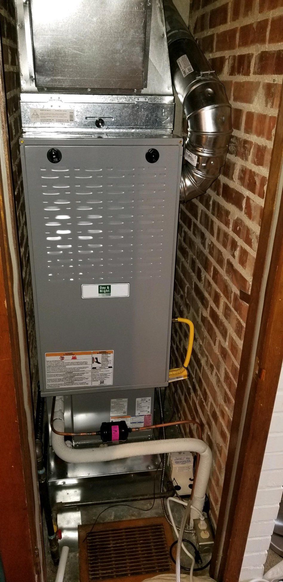 Furnace unit in a brick-lined alcove. Metallic gray and silver with yellow gas line and white PVC pipes.
